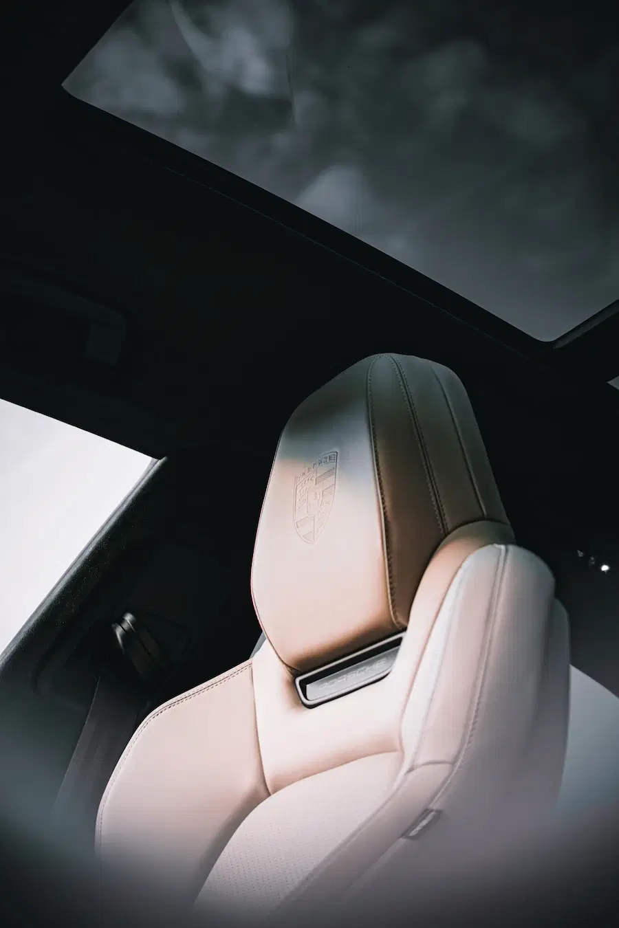 Luxurious beige Porsche seat with logo embossing beneath a panoramic glass roof, highlighting premium interior design.